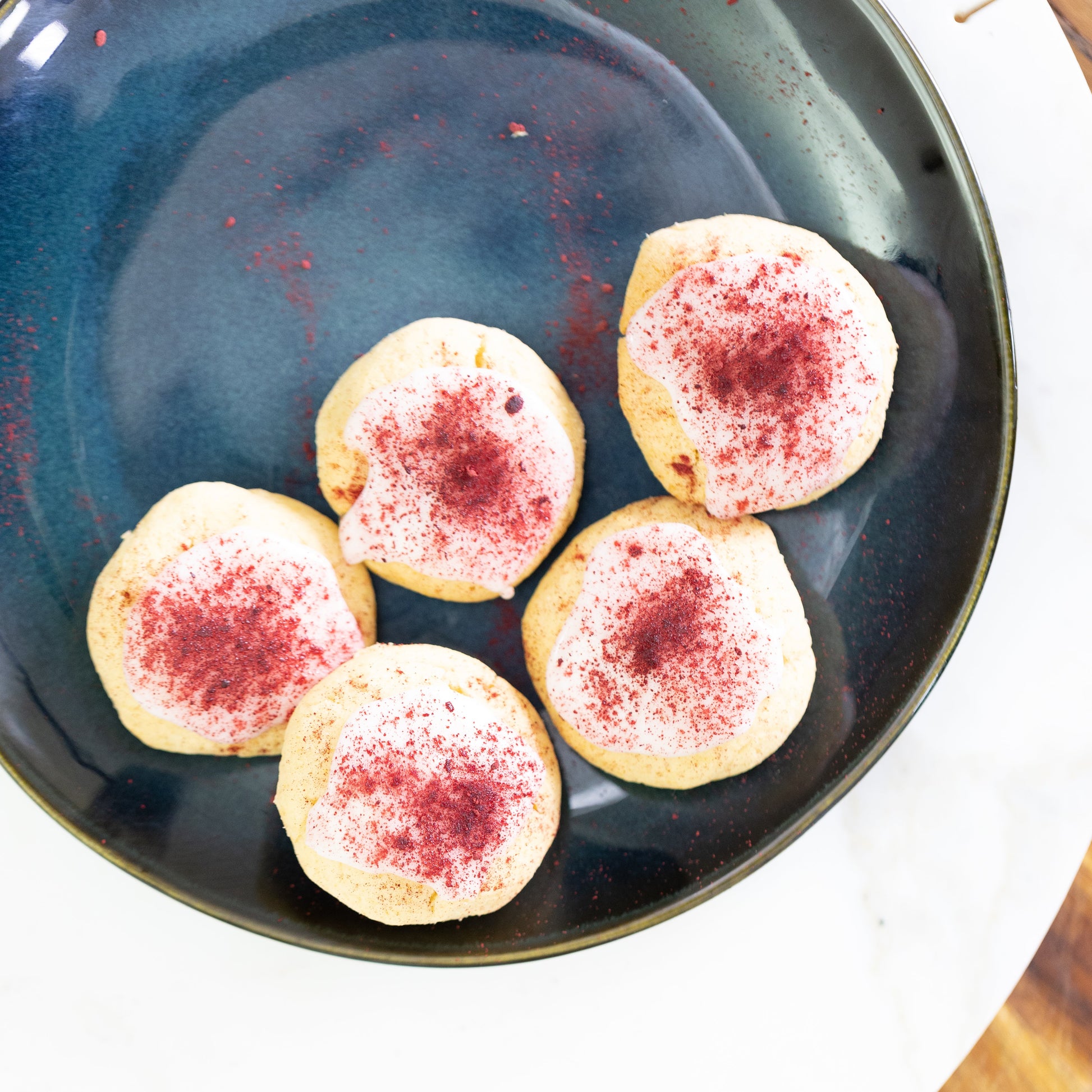 Five cookies with red sprinkles on a dark blue plate, placed on a wooden surface.